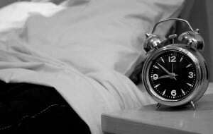Image shows an alarm clock next to a bed, in black and white.