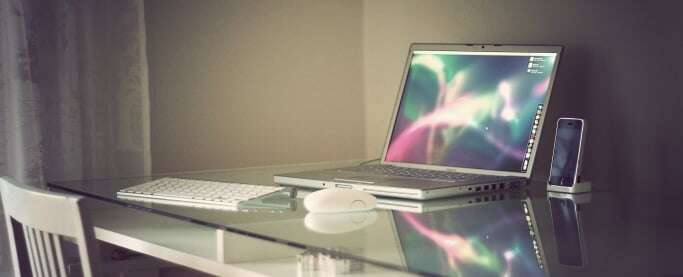 Image shows an Apple laptop on a glass desk. 