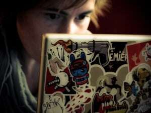Image shows a woman writing at a laptop, the back of which is covered in stickers.