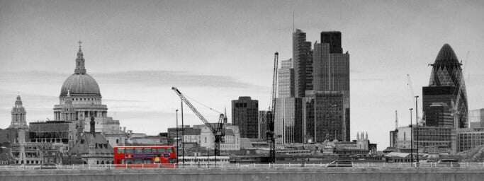 Image shows the London skyline in black and white, with a London bus in front of it in red.