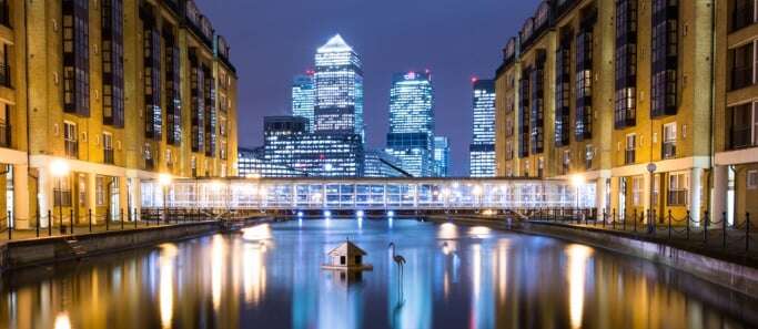 Image shows Canary Wharf at night, seen from between two rows of buildings over a canal.