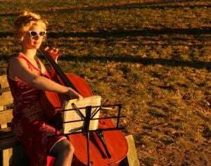 Image shows a woman wearing sunglasses playing cello in a park at sunset.