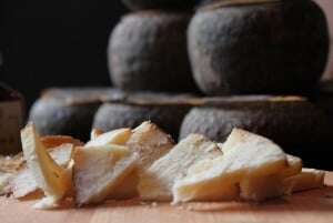 Image shows cut cheese in the foreground on a table, with wheels of cheese in black wax stacked in the background.