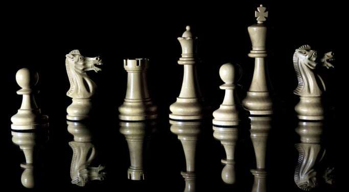 Image shows a row of chess pieces against a black background and reflected on the surface of a black table.