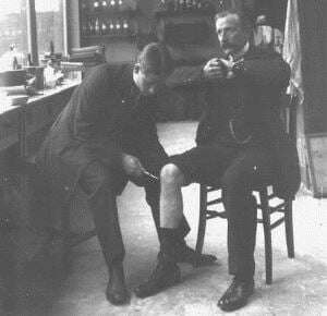 Image shows an old black-and-white photograph of two men, both seated. One is tapping the other's knee with a hammer, while he holds his arms in the Jendrassik manoeuvre. 
