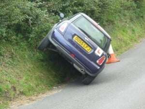 Image shows a car with a learner sticker on the back, crashed into the verge beside a road.