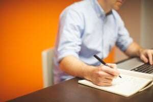 Image shows someone making notes while working at a laptop.