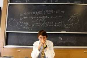 Image shows a boy in a lab coat in front of a blackboard full of formulae.