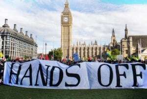 Image shows protesters in front of Parliament with a sign that reads "hands off".