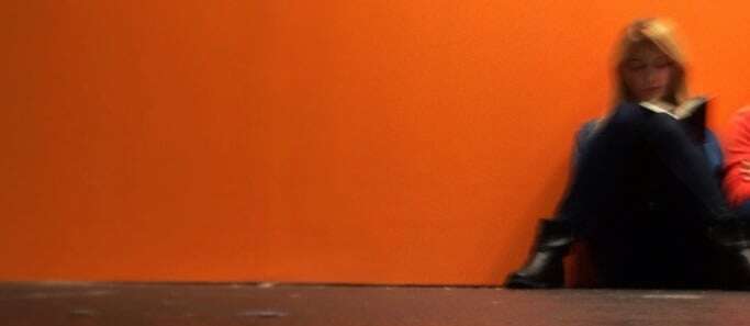 Image shows a woman reading a book, leading against an orange wall.