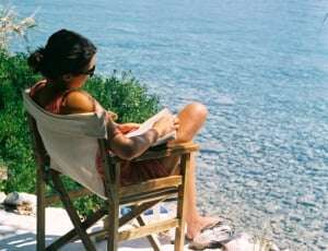 Image shows a young woman sitting on a chair and reading a book, looking out over a clear blue sea.