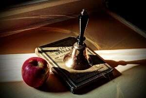 Image shows a bell, an apple and a spelling book on a wooden table.