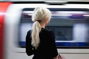 Image shows a young woman standing on a platform in front of a moving London Underground train.