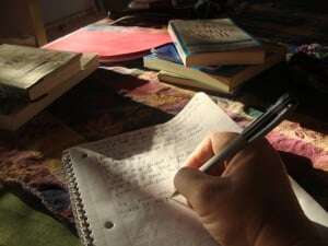 Image shows someone writing in a notebook, with stacks of books on their desk.