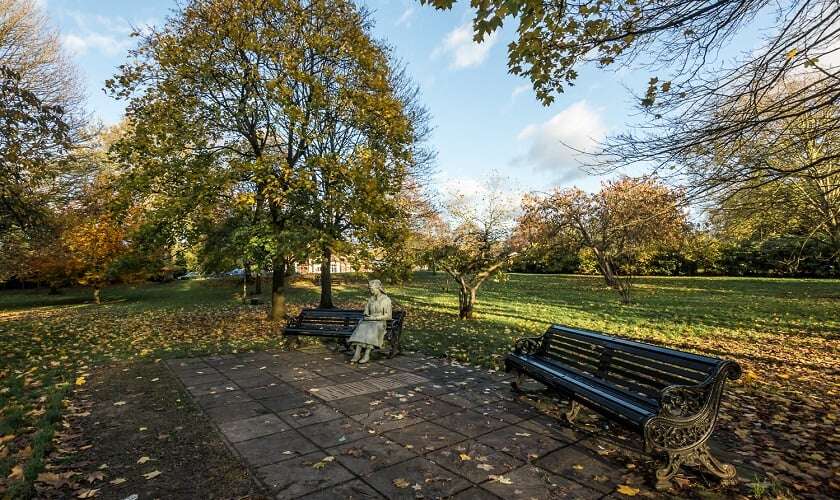 Image shows a park, with a row of benches, and a statue of Vera Brittain sitting on one of them.